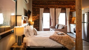 A modern hotel room with a brick wall, a bed with white linens, a wooden nightstand, a lamp, and a view of the kitchen. - Studio Suite Yadkin Valley Downtown Elkin (Elkin)