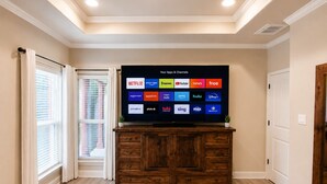 Un televisor mostrando diversas aplicaciones y canales de transmisión, un mueble de madera debajo, y una ventana con persianas a la izquierda.