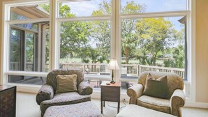 A spacious room with large windows offering a view of trees and a deck. There are two armchairs and a small table with a lamp. - 4 Minute Walk to Beach!  Private Pool With Pool Heater.*  Back Deck w/ Lagoon & Golf Course Views! (Hilton Head Island)
