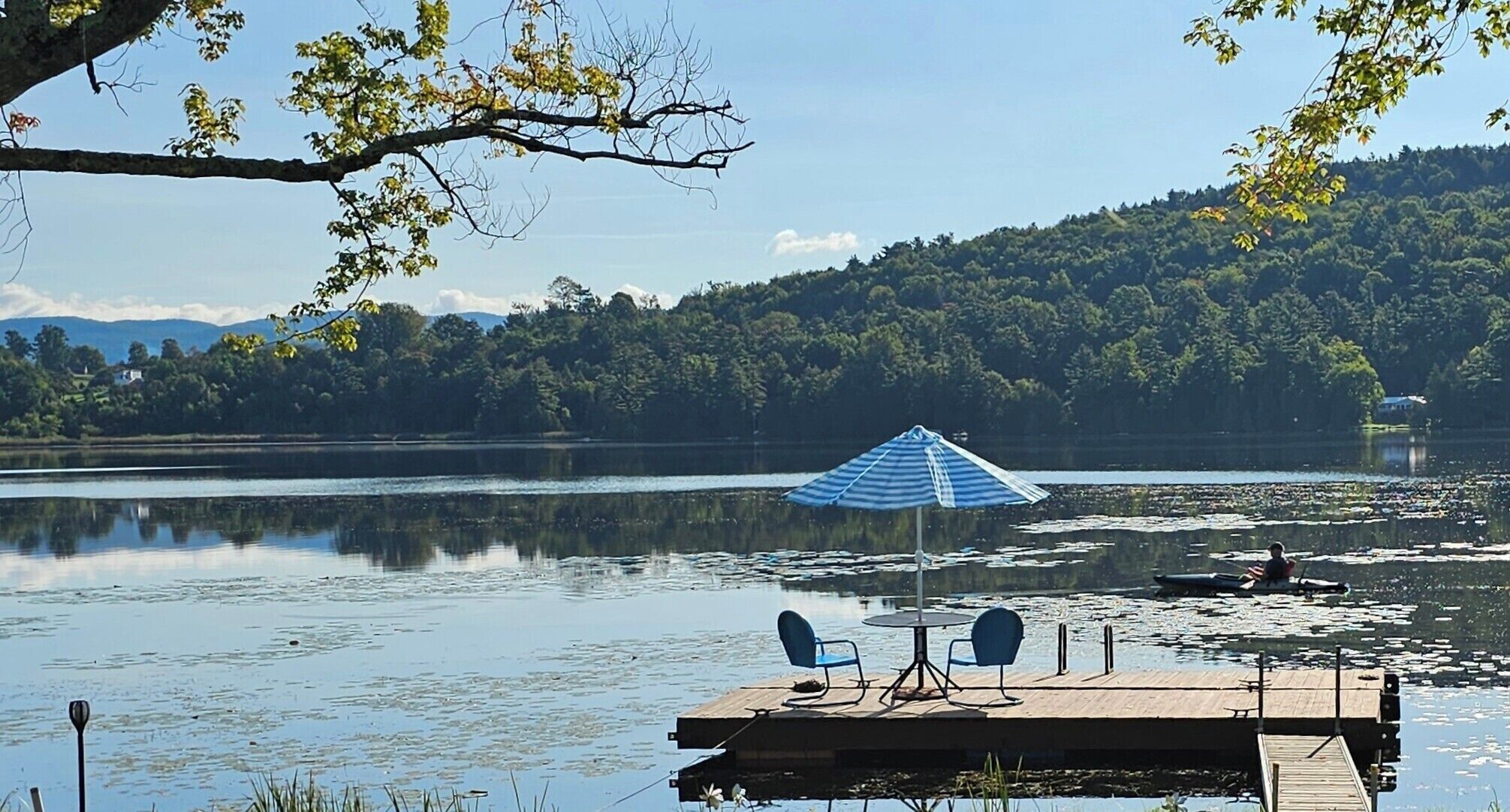 Une jetée au bord du lac, avec un parasol bleu, deux chaises et une table.