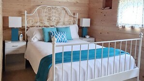 Un dormitori de cabana de fusta amb un llit de metall blanc, un ventilador de sostre i una petita finestra amb cortines.