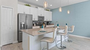 A modern kitchen with white cabinets, stainless steel appliances, a central island with bar stools, and pendant lighting. - Lovely Family Home - 8 Min to Disney! (Kissimmee)