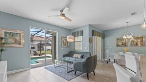 A spacious living area with a grey sofa, a glass coffee table, a dining table with chairs, a ceiling fan, and a view of a swimming pool. - Lovely Family Home - 8 Min to Disney! (Kissimmee)