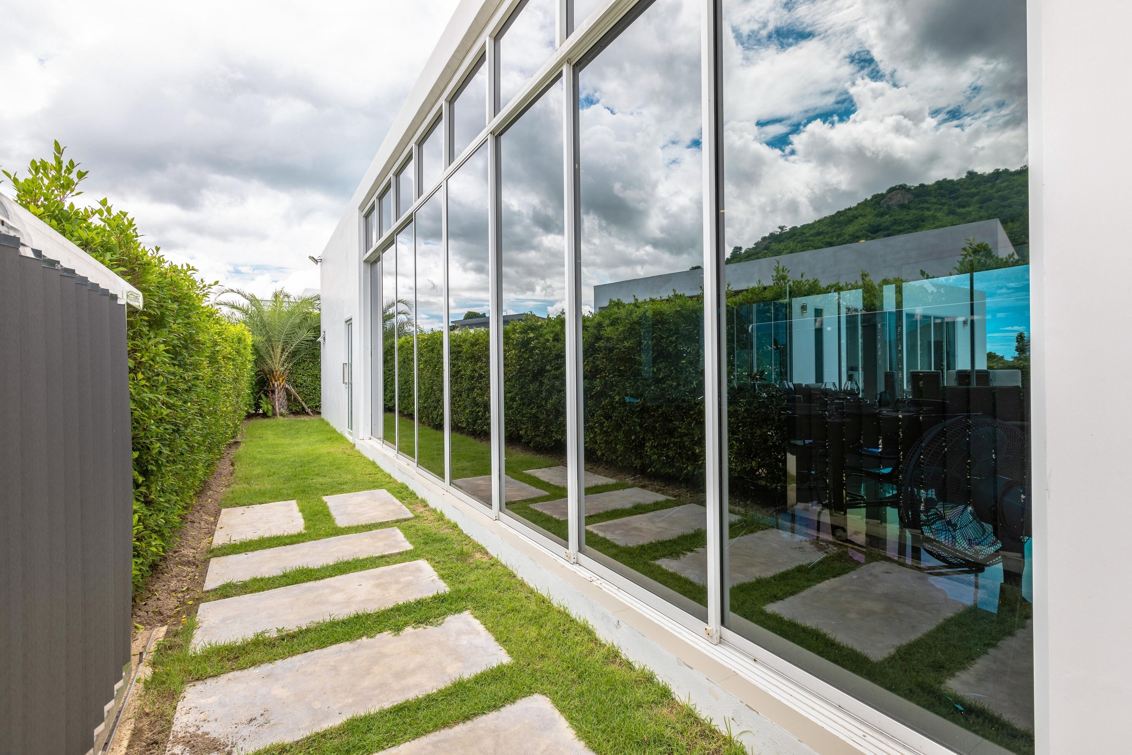 A modern building with large glass windows, a stone pathway, and a view of a lush garden and distant hills.