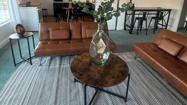 Un salon avec un canapé en cuir marron, une table basse ronde sur laquelle est posé un vase, et une cuisine en arrière-plan.