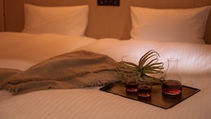 Un letto rifatto con cura, lenzuola bianche, una coperta marrone e un vassoio con due bicchieri di liquido rosso e una bottiglia di vetro trasparente.
