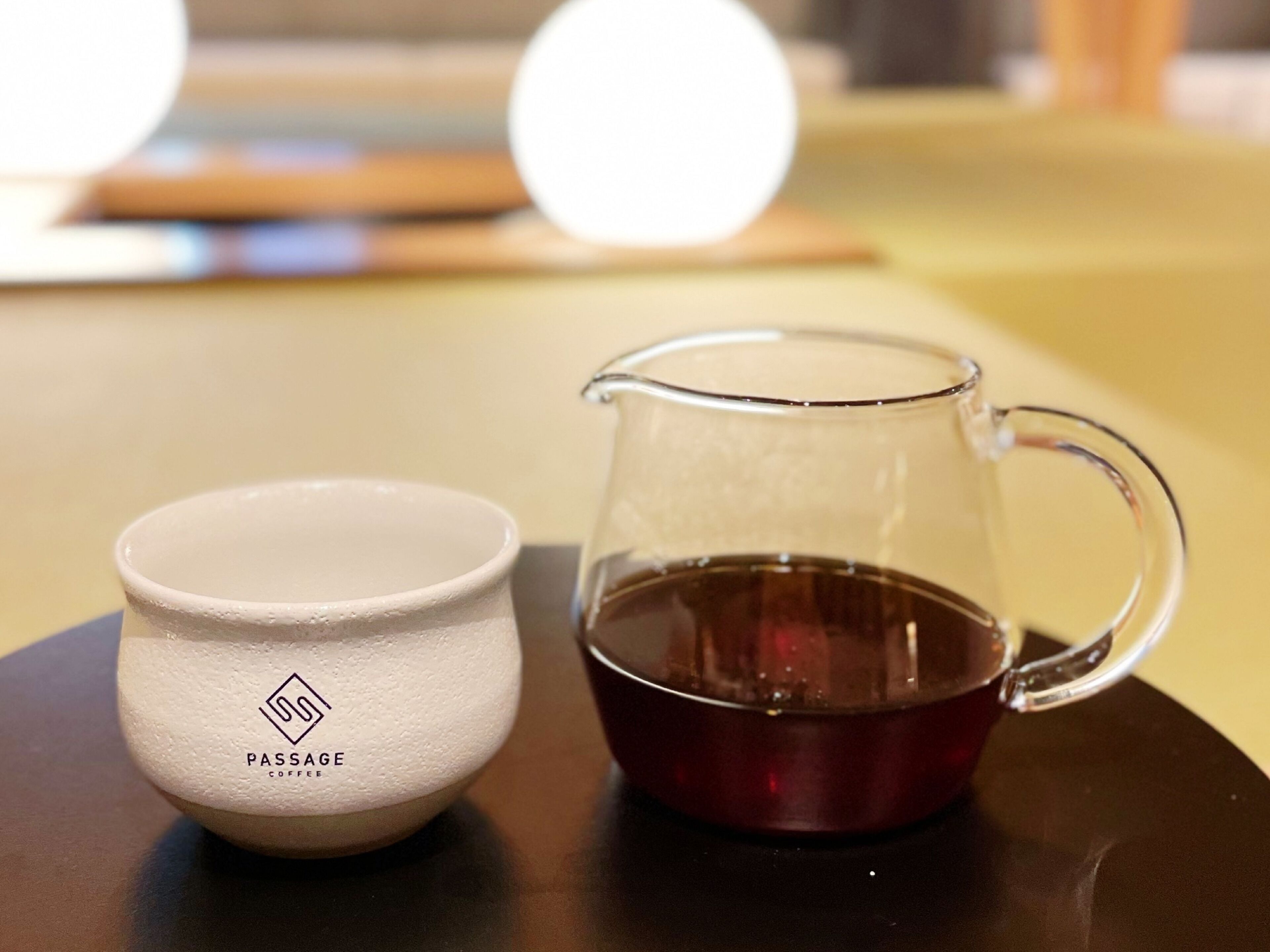 A white ceramic cup with "PASSAGE COFFEE" text and a clear glass pitcher with a handle, both on a dark surface.
