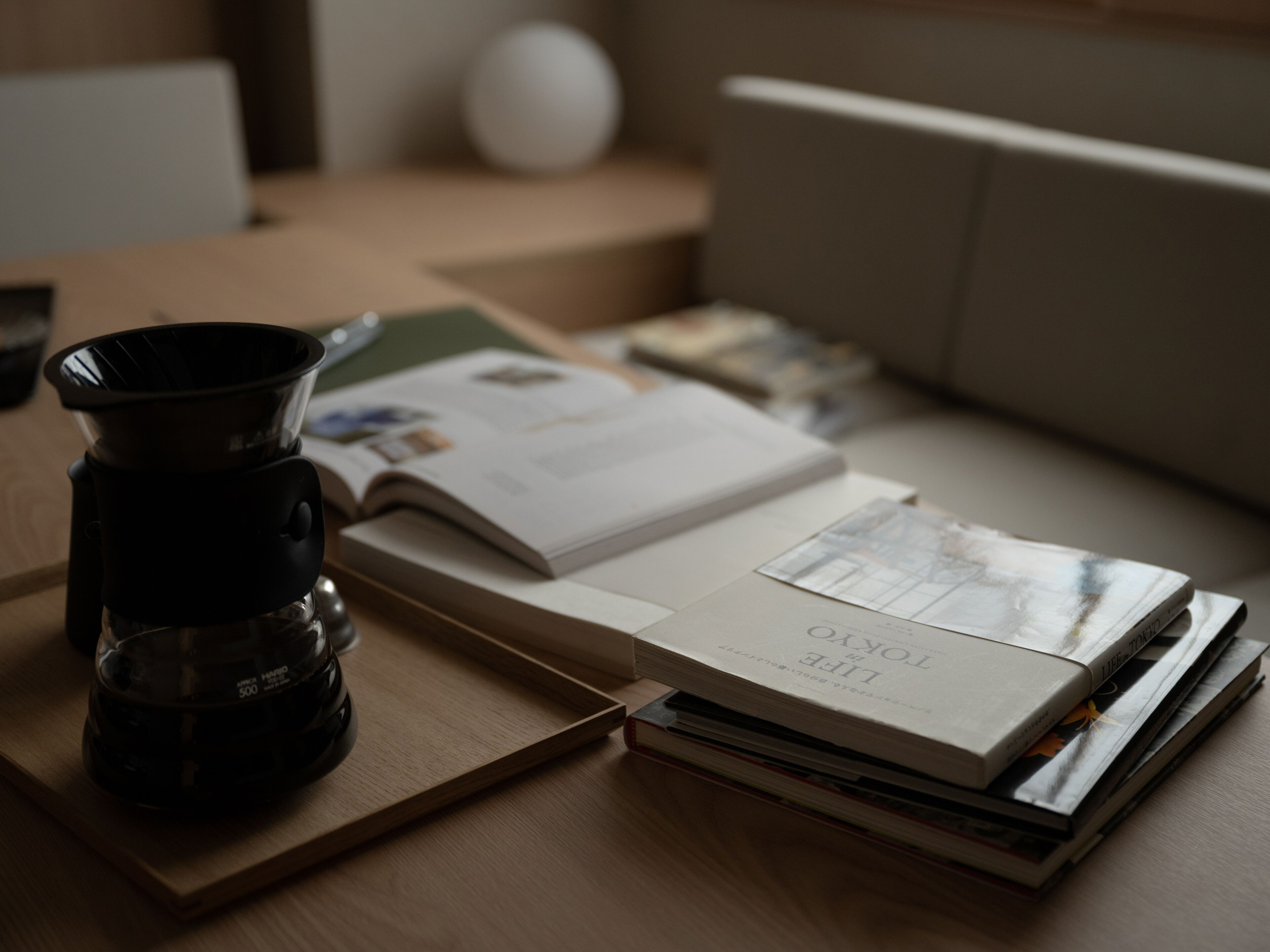 A coffee maker, an open book, and a stack of magazines on a wooden table.