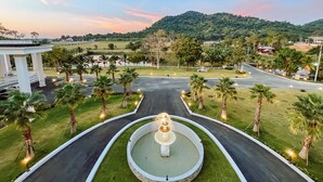 A landscaped area with a fountain, palm trees, and a road leading to a building with a white facade. - Grand Lord Hotel& Residence (Sattahip)