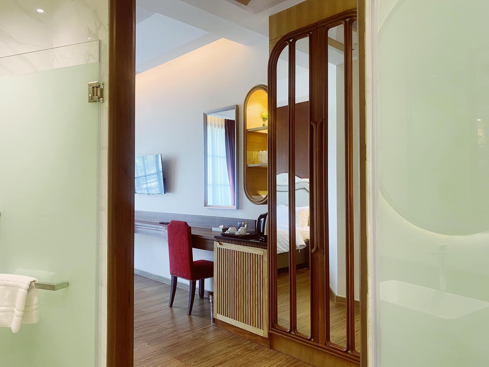 Habitación de hotel con un armario de madera, un escritorio con silla, una cama, un televisor montado en la pared y un baño con puerta de cristal.