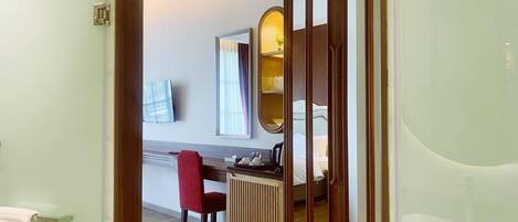 Habitación de hotel con un armario de madera, un escritorio con silla, una cama, un televisor montado en la pared y un baño con puerta de cristal.