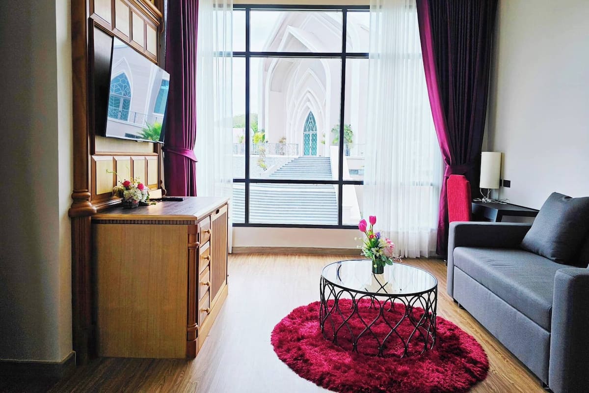 A modern living room with a large window, a television, a sofa, a coffee table, and a desk with a vase of flowers.