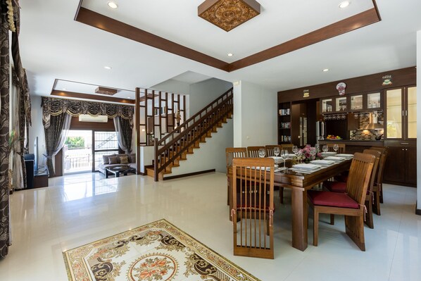 A spacious living area with a wooden staircase, a dining table set for six, and a kitchen area with wooden cabinets and a coffee station.