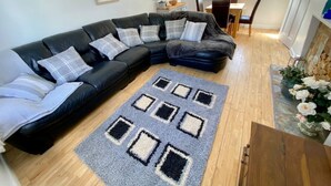 A living room with a black sectional sofa, a patterned rug, and a dining area with a table and chairs. - A beautiful refurbished Cornish Cottage with BBQ & Patio (Falmouth)