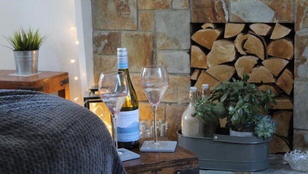 A cozy living room with a stone fireplace, a wooden table with wine bottles and glasses, and a potted plant. - A beautiful refurbished Cornish Cottage with BBQ & Patio (Falmouth)
