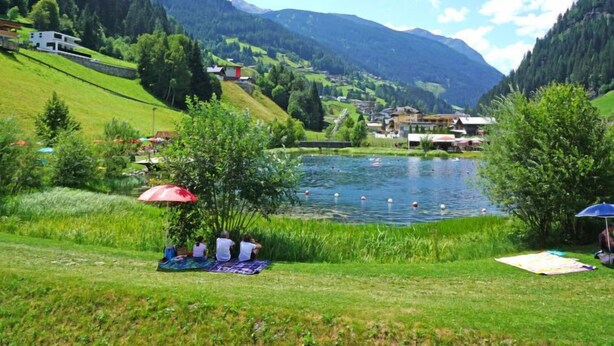 Blick auf einen See mit einer Rasenfläche, einem Picknickplatz und Gebäuden im Hintergrund.