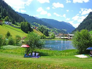 Vista para um lago com área gramada, mesa de piquenique e edifícios ao fundo.