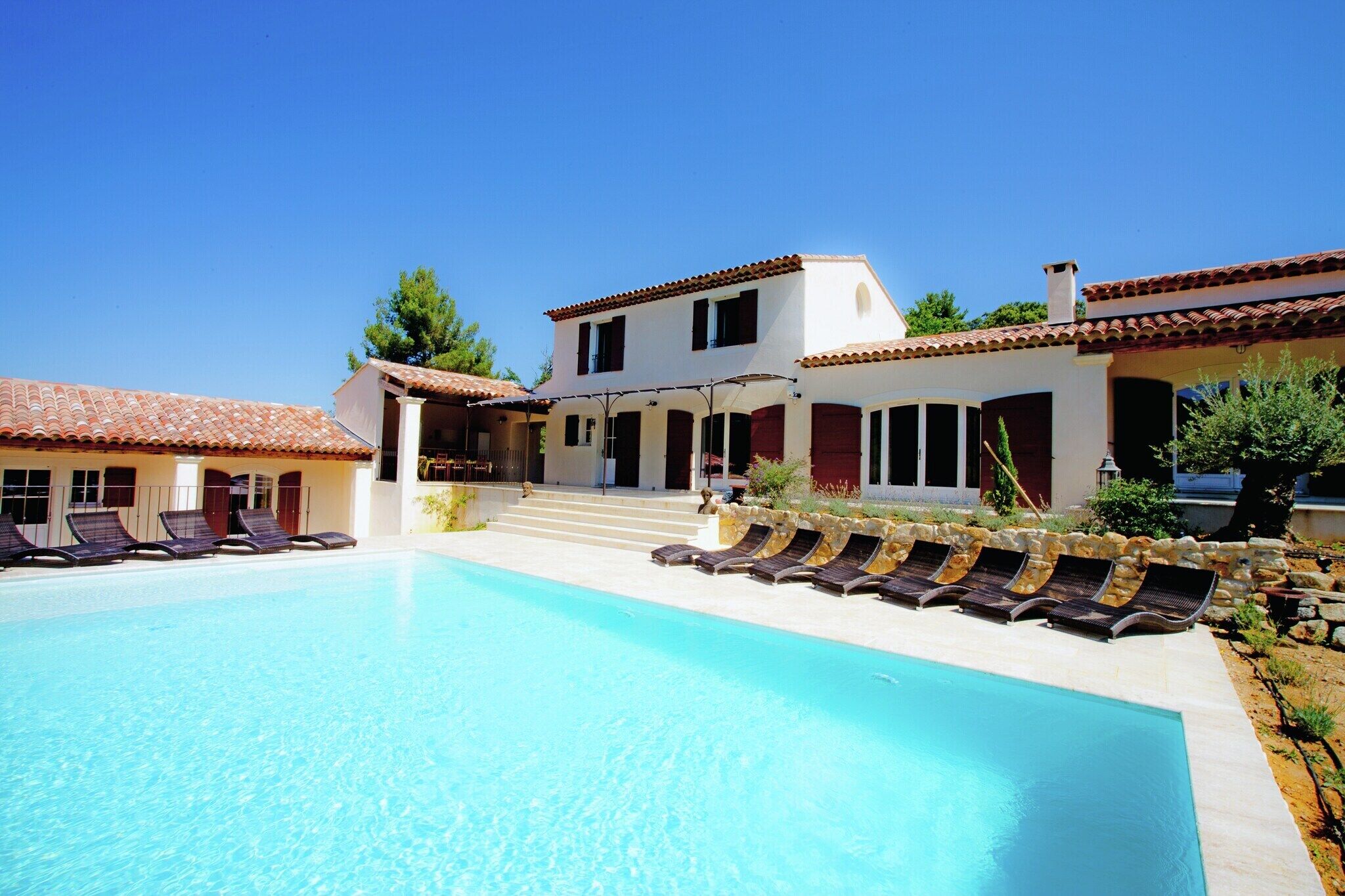 A poolside view of a resort with a clear blue pool, sun loungers, and a two-story building with a tiled roof.