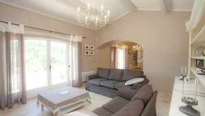 A spacious living room with a large sofa, a coffee table, and a chandelier. There is a sliding glass door leading to a balcony. - Villa in Provence with Infinity Pool (Apt)