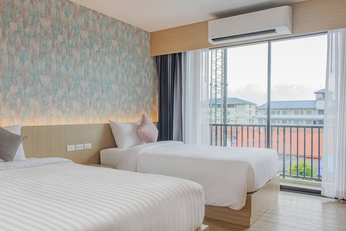Habitación de hotel con dos camas, un ventanal con vistas a edificios y un equipo de aire acondicionado.