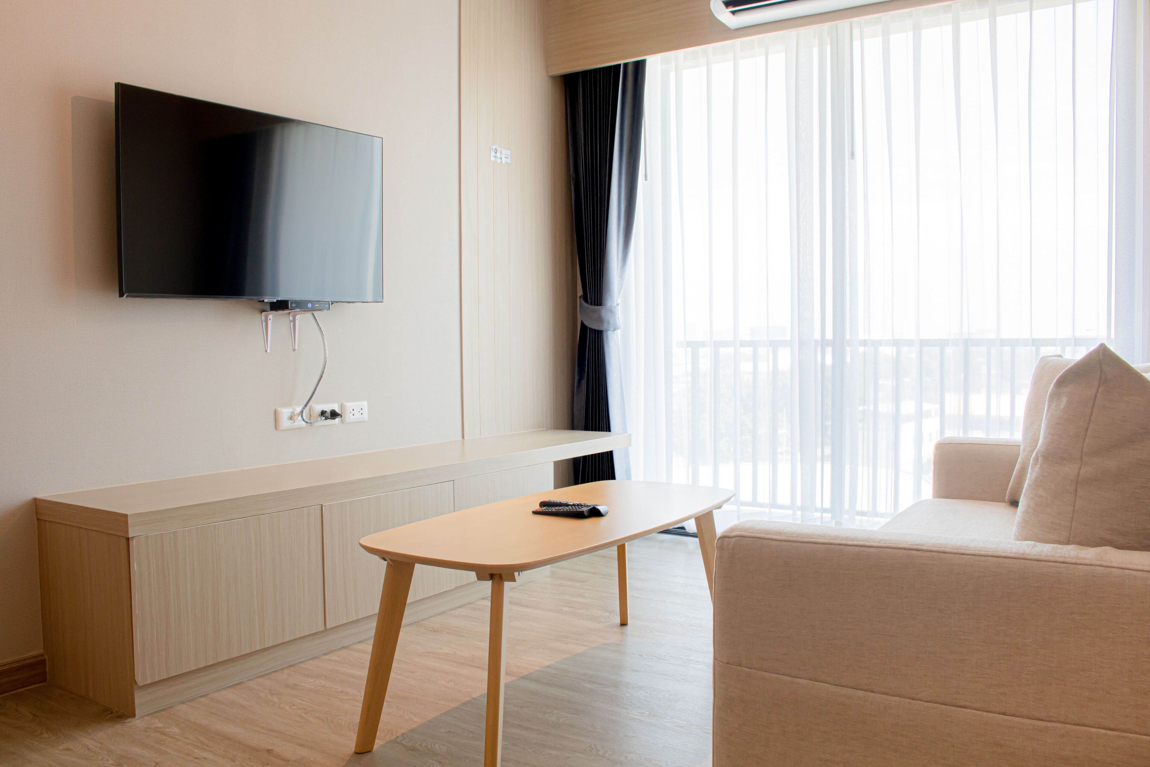 A modern living room with a flat-screen TV mounted on the wall, a wooden coffee table, a beige sofa, and a large window with sheer curtains.