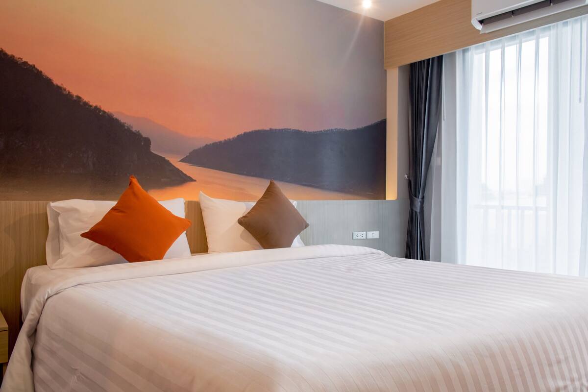 Habitación de hotel con cama, almohadas, mesita de noche, equipo de aire acondicionado, ventana con cortinas y un mural en la pared que representa un atardecer sobre el agua.