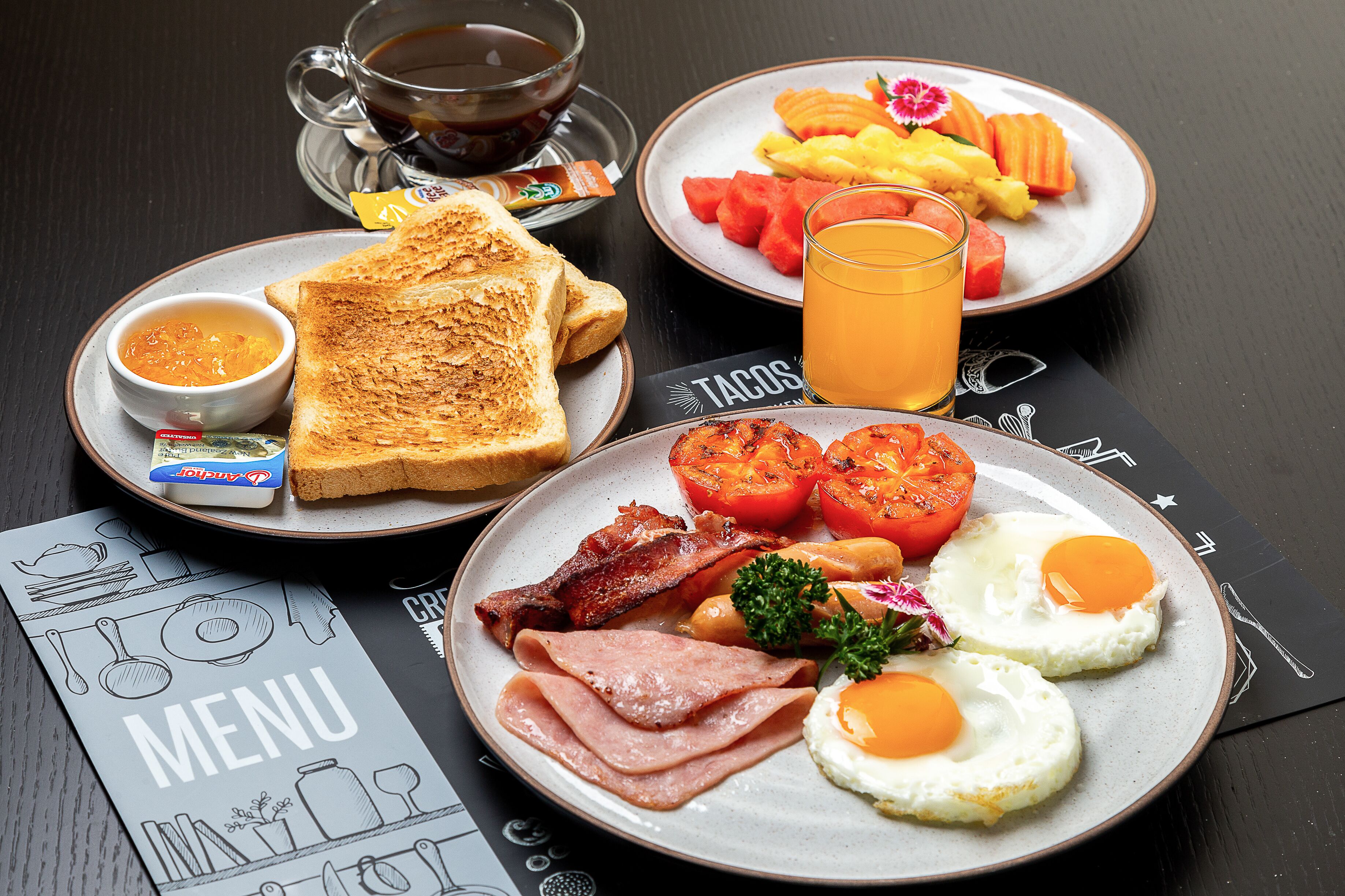 Una mesa con una variedad de alimentos para el desayuno, incluyendo tostadas, huevos, tocino, tomates y un vaso de jugo de naranja.