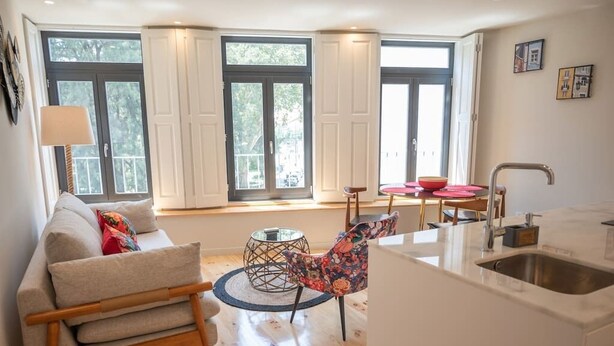 Un salon moderne avec un canapé, une table basse ronde et un fauteuil. On y trouve également un coin cuisine avec un évier et une fenêtre donnant sur des arbres.