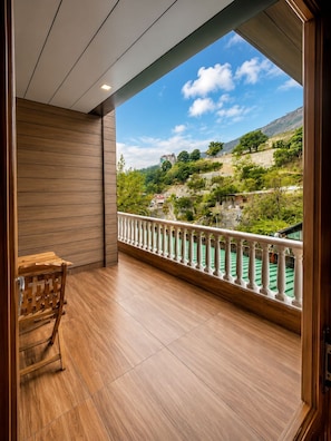 A balcony with a wooden floor, a white balustrade, and a view of a green landscape with trees and a building in the distance.