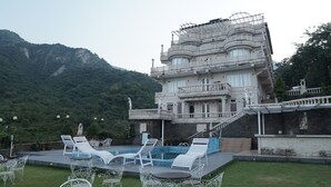 Um edifício grande com área de piscina, mobiliário ao ar livre e montanhas ao fundo.