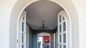 A hallway with a dark floor, white walls, and an arched doorway leading to a room with a red wall and a chandelier.