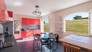 A modern kitchen with red cabinets, a dining area with a wooden table and chairs, and a view of a grassy field through large windows.