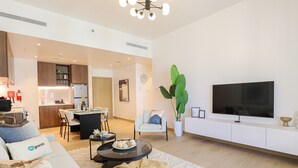 A modern living room with a sofa, coffee table, and a TV. There is a dining area with a table and chairs, and a kitchen in the background. - HiGuests - La Cote B2 (Dubai)