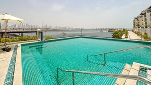 A swimming pool with clear blue water, surrounded by a tiled deck and a view of a city skyline across a body of water. - HiGuests - La Cote B2 (Dubai)