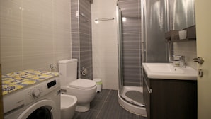 A compact bathroom with a washing machine, toilet, and a glass-enclosed shower. - Albania Dream Beachfront Apartment (Sarandë)