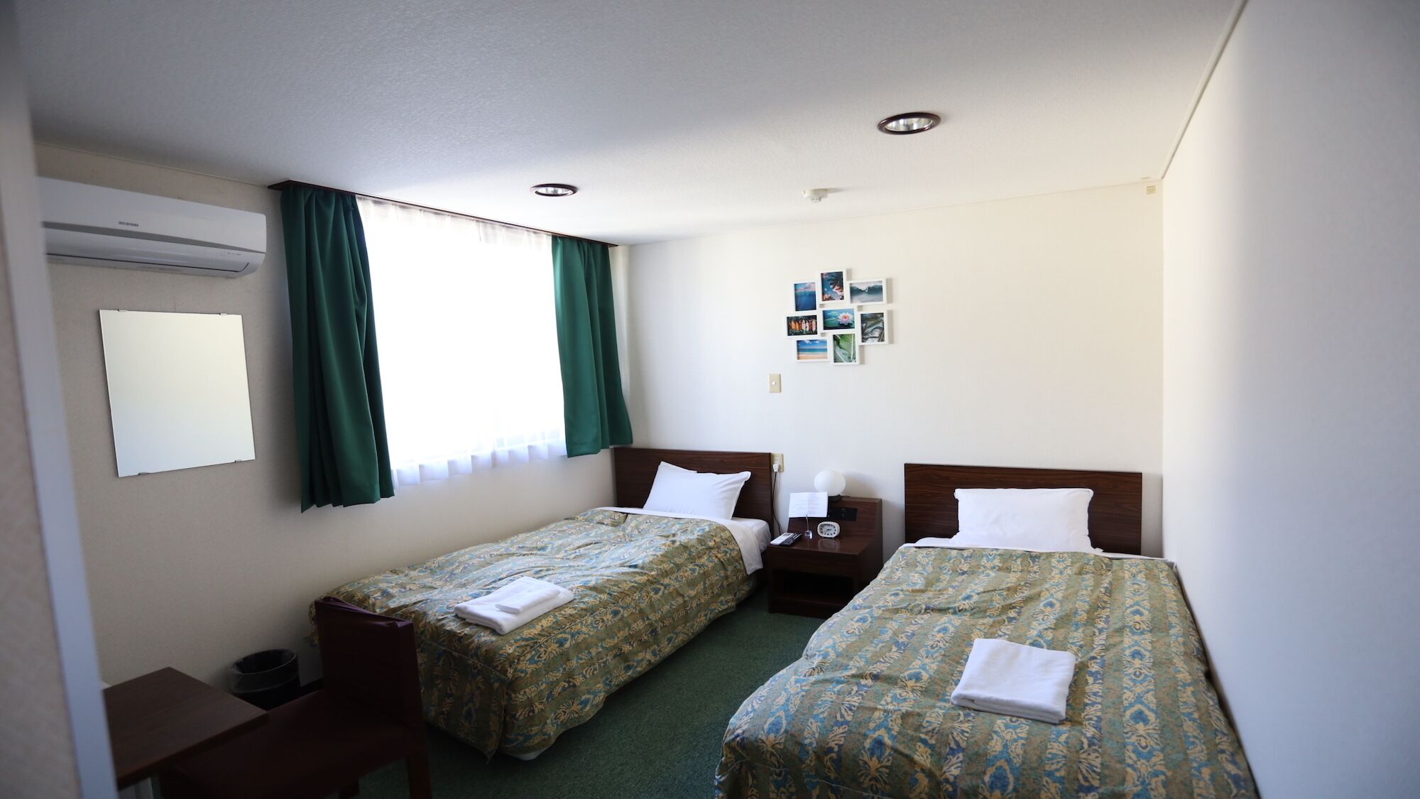 A hotel room with two beds, a desk, a chair, a small table, a wall-mounted air conditioner, and a window with curtains.
