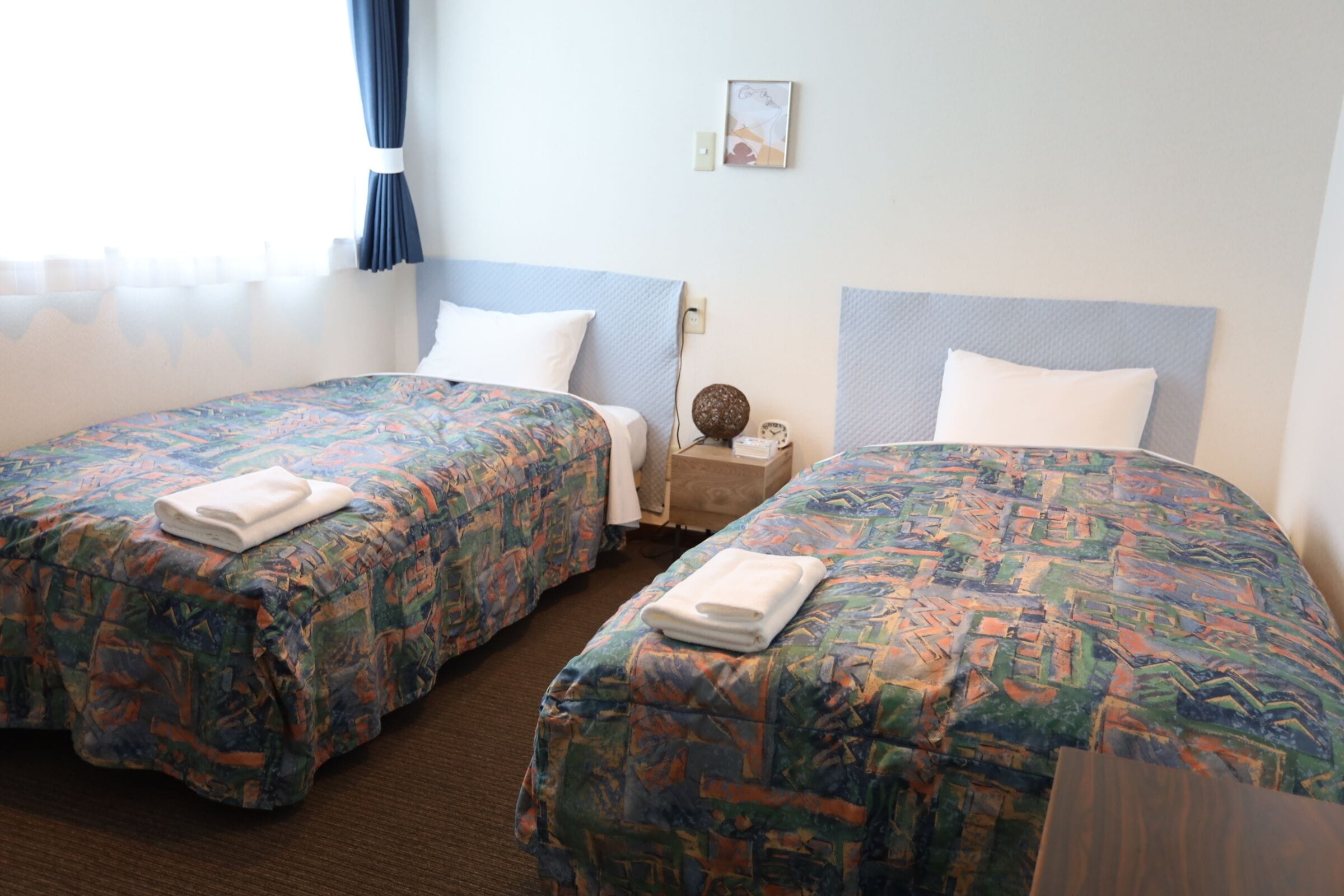 A hotel room with two single beds, each with a patterned bedspread and white pillows. There is a wooden nightstand between the beds with a lamp and a small decorative item. The room has a simple, clean design with a light-colored wall and a window with curtains.