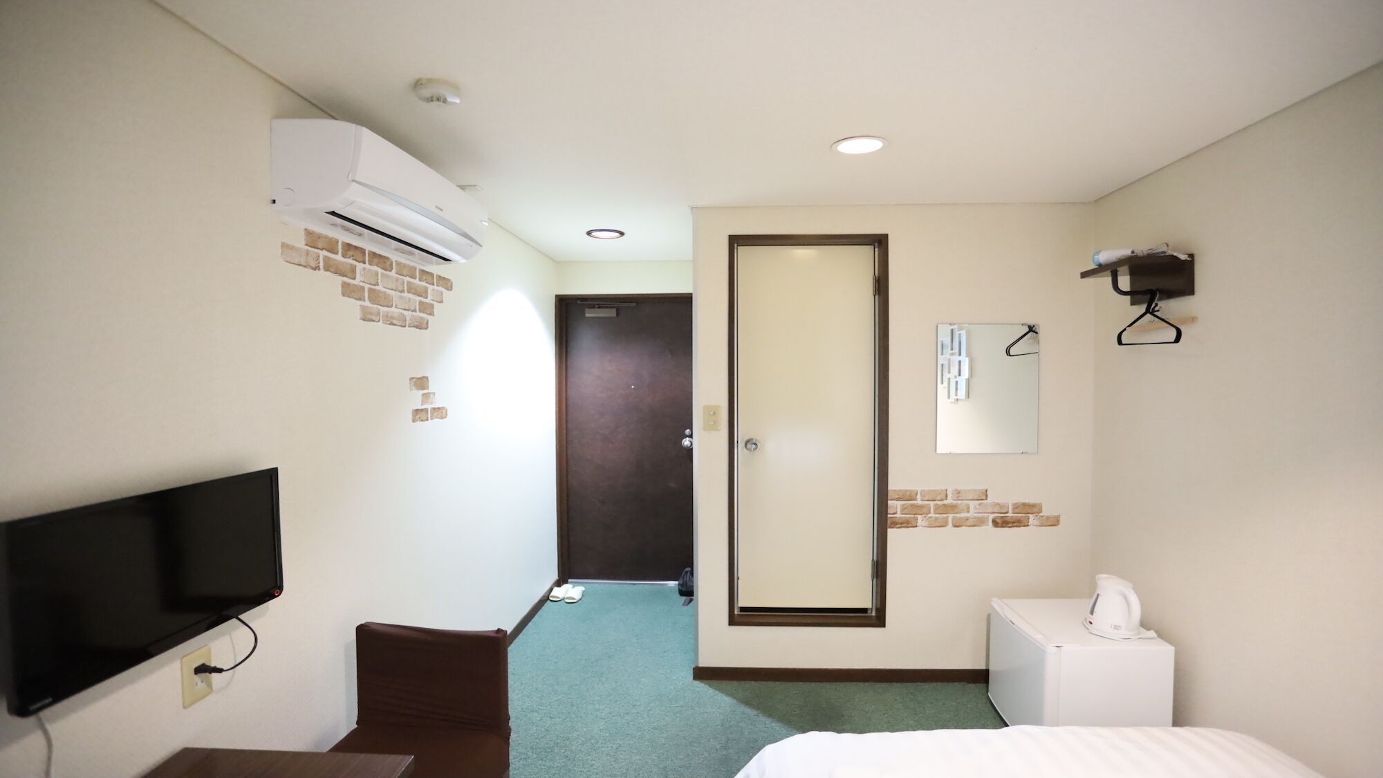 A hotel room with a flat-screen TV, a bed, a small table, a mirror, and a wall-mounted heater.