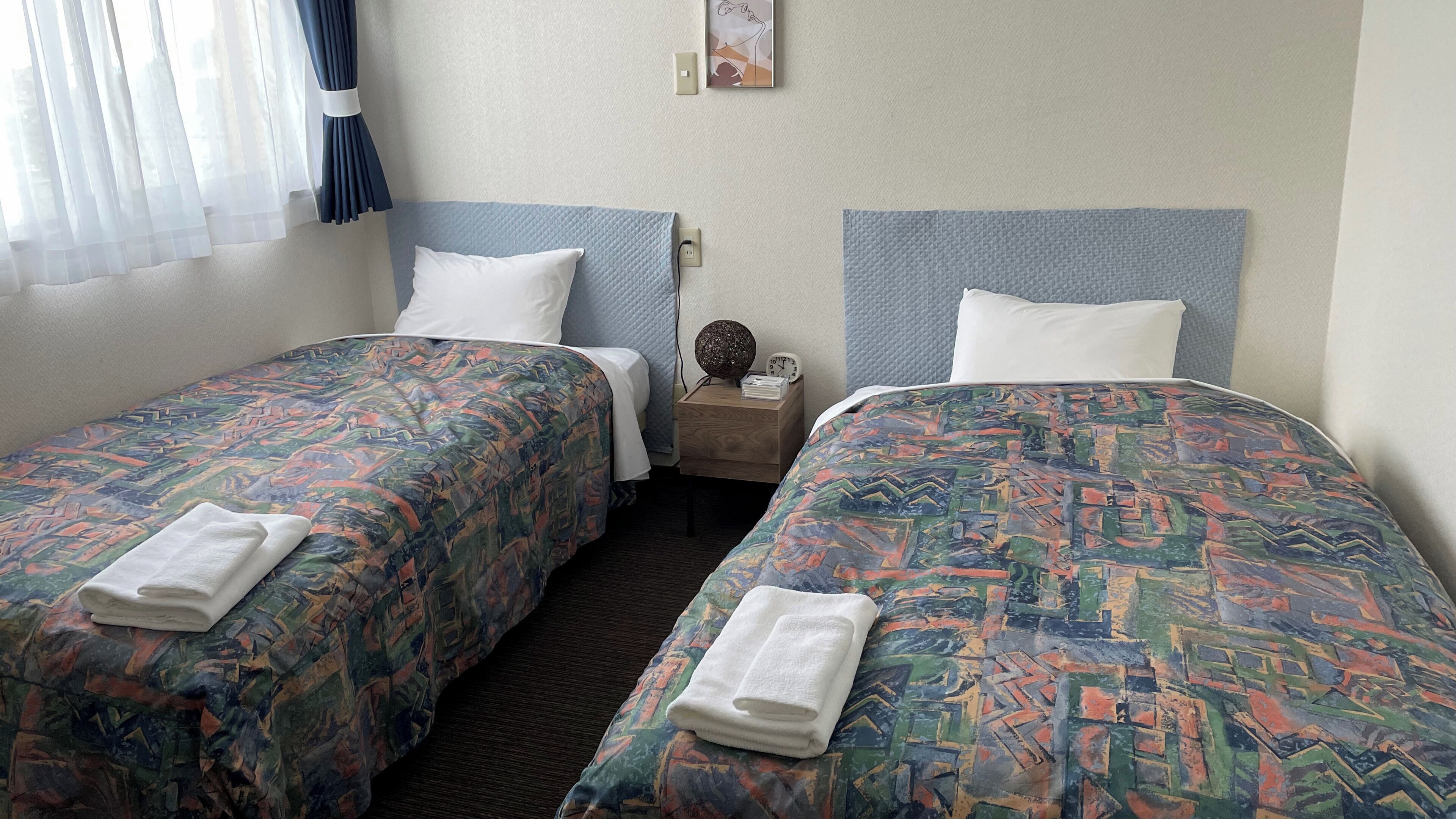 A hotel room with two beds, each with a patterned bedspread, white pillows, and a small bedside table with a clock and a decorative item.