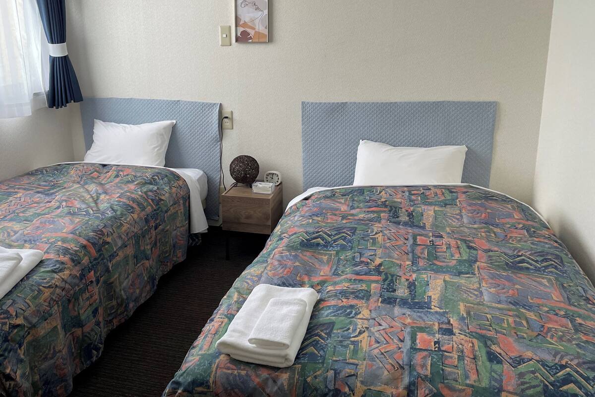 A hotel room with two beds, each with a patterned bedspread, white pillows, and a small bedside table with a clock and a decorative item.
