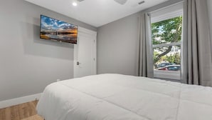 A bedroom with a bed, a TV mounted on the wall, a window with curtains, and a view of the outside.