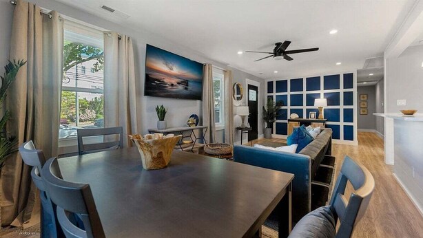A modern living room with a dining area, a ceiling fan, a large window with curtains, and a blue accent wall.