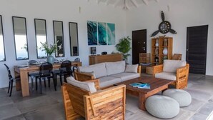 Ein modernes Wohnzimmer mit Couch, Esstisch und Stühlen. Ein Holztisch, ein Bücherregal mit Dekoration und eine große Wanduhr sind ebenfalls zu sehen.