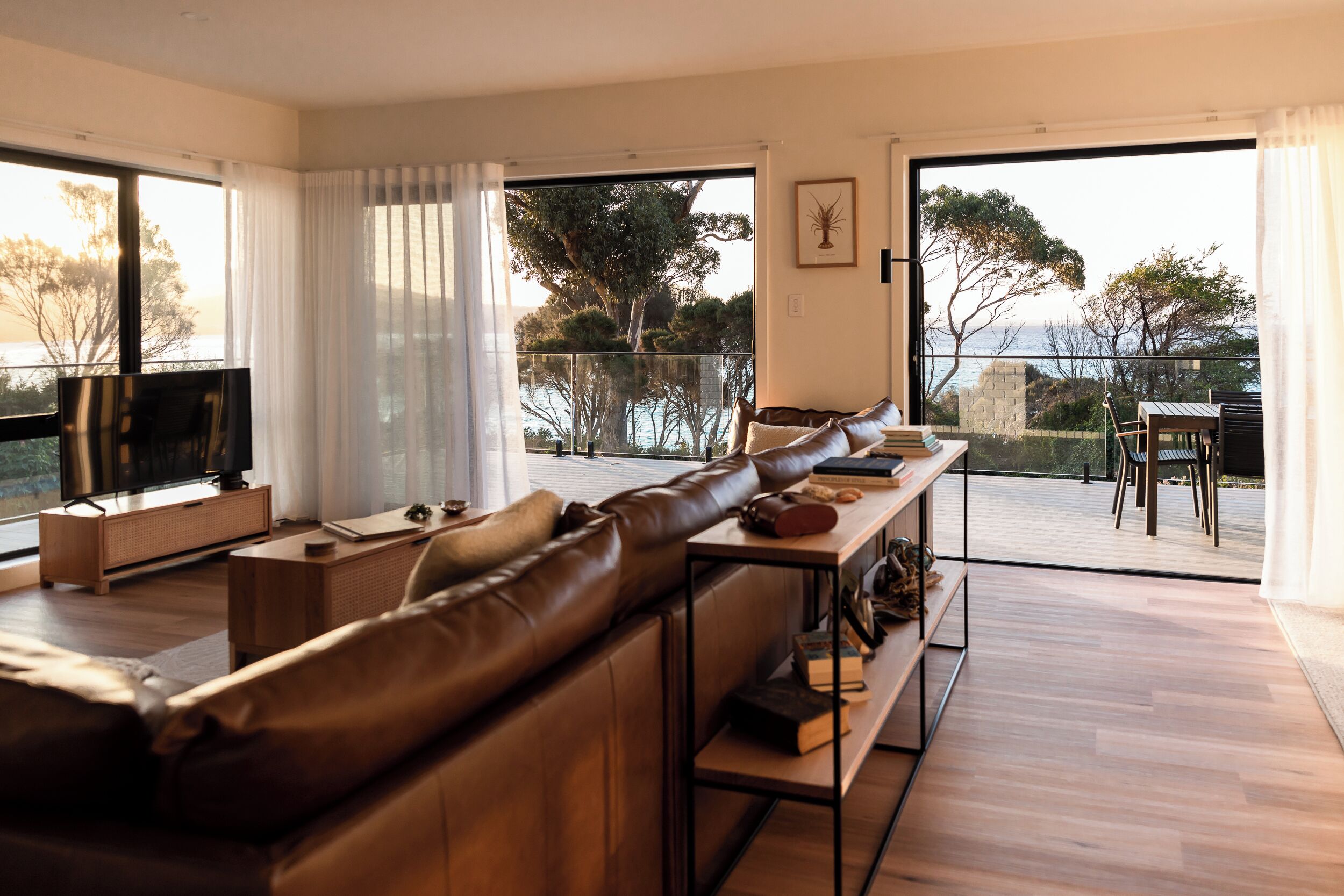 A modern living room with a leather sofa, a flat-screen TV, and a wooden coffee table. There is a balcony with a view of the sea and trees.