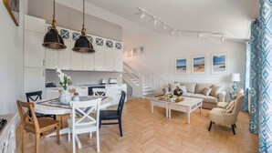 Una cocina moderna con armarios blancos, un comedor con mesa y sillas de madera, una sala de estar con sofá y mesa de centro, y una escalera que lleva a un nivel superior.