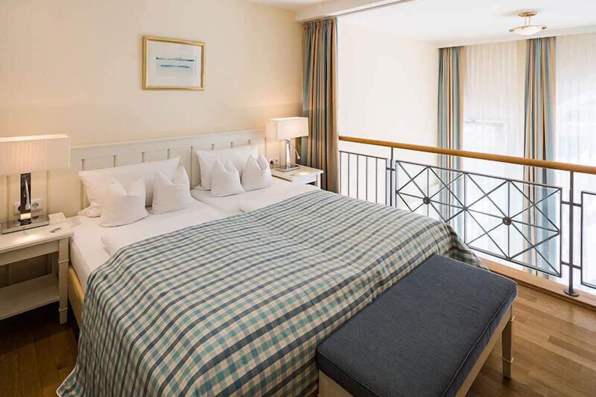 Habitación de hotel con una cama grande, una mesita de noche con lámpara, un banco, una ventana con cortinas y una barandilla.