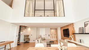 Un espacio habitable amplio con suelo de madera, un sofá, una mesa de comedor con adornos, un televisor montado en la pared y una escalera que sube a un nivel superior.
