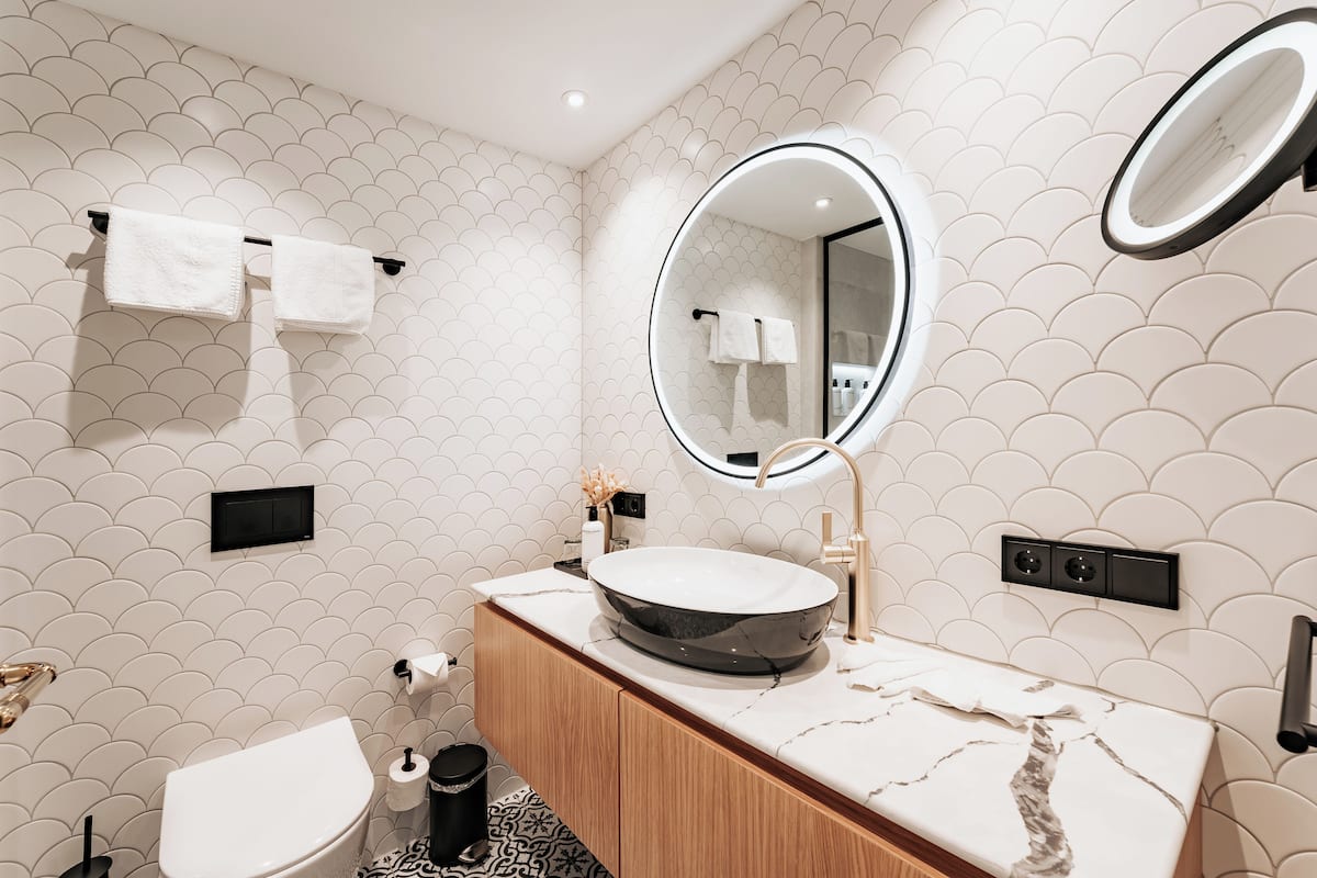Un baño moderno con un espejo redondo, paredes de azulejos blancos, un mueble de tocador de madera y un inodoro.