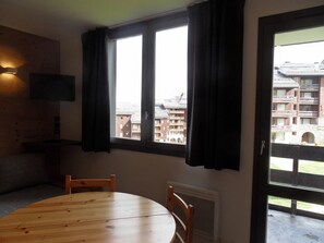 Een kamer met een ronde houten tafel, twee stoelen, een flatscreen televisie aan de muur en een raam met donkere gordijnen.