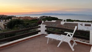 Una terrazza con due poltrone bianche, pavimento piastrellato e vista sul mare e sulle montagne all'orizzonte durante il tramonto.